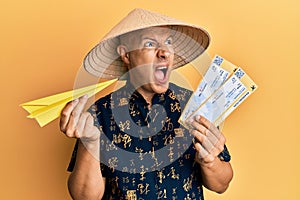 Middle age bald man wearing traditional asian hat holding boarding pass angry and mad screaming frustrated and furious, shouting