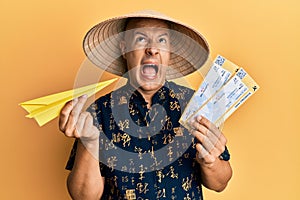 Middle age bald man wearing traditional asian hat holding boarding pass angry and mad screaming frustrated and furious, shouting