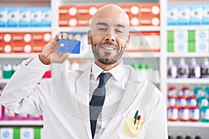 Middle age bald man working at pharmacy drugstore holding credit card looking positive and happy standing and smiling with a
