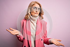 Middle age beautiful blonde business woman wearing elegant pink jacket and glasses clueless and confused expression with arms and