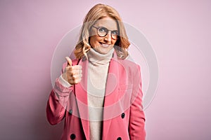 Middle age beautiful blonde business woman wearing elegant pink jacket and glasses doing happy thumbs up gesture with hand