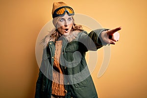 Middle age beautiful blonde skier woman wearing snow sportwear and ski goggles Pointing with finger surprised ahead, open mouth