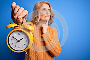 Middle age beautiful blonde woman holding vintage alarm clock over blue background serious face thinking about question, very