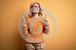 Middle age beautiful blonde woman wearing casual sweater and glasses over yellow background Pointing up looking sad and upset,