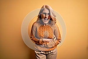 Middle age beautiful blonde woman wearing casual sweater and glasses over yellow background smiling and laughing hard out loud