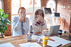 Middle age beautiful business workers working together using laptop at the office shocked covering mouth with hands for mistake