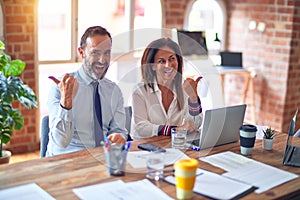 Middle age beautiful business workers working together using laptop at the office smiling with happy face looking and pointing to