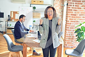 Middle age beautiful businesswoman wearing jacket and glasses standing at the office celebrating surprised and amazed for success