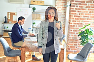 Middle age beautiful businesswoman wearing jacket and glasses standing at the office doing happy thumbs up gesture with hand