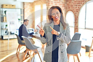 Middle age beautiful businesswoman wearing jacket and glasses standing at the office smiling and looking at the camera pointing