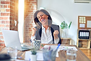 Middle age beautiful businesswoman working using laptop at the office very happy and excited doing winner gesture with arms