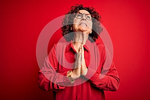 Middle age beautiful curly hair woman wearing casual shirt and glasses over red background begging and praying with hands together