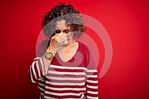 Middle age beautiful curly hair woman wearing casual striped sweater over red background tired rubbing nose and eyes feeling