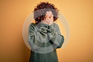 Middle age beautiful curly hair woman wearing casual sweater over  yellow background with sad expression covering face