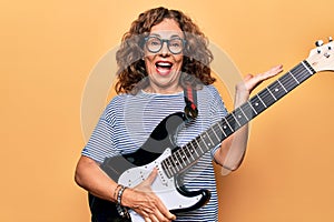 Middle age beautiful musician woman playing electric guitar over isolated yellow background celebrating achievement with happy