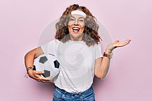 Middle age beautiful sporty woman playing soccer holding football bal over pink background celebrating achievement with happy
