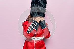 Middle age beautiful wales guard woman wearing traditional uniform over pink background with sad expression covering face with