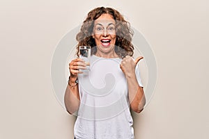 Middle age beautiful woman drinking glass of water to refreshment over white background pointing thumb up to the side smiling