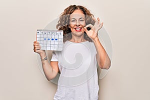 Middle age beautiful woman holding climatology calendar showing cloudy and rainy weather doing ok sign with fingers, smiling photo