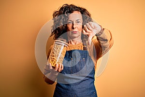 Middle age beautiful woman holding jar with macaroni pasta over isolated yellow background with angry face, negative sign showing