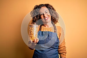 Middle age beautiful woman holding jar with macaroni pasta over isolated yellow background with a happy face standing and smiling
