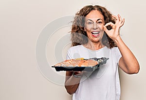 Middle age beautiful woman holding takeaway tray with sushi over isolated white background doing ok sign with fingers, smiling
