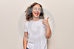 Middle age beautiful woman listening to music using headphones over white background pointing thumb up to the side smiling happy