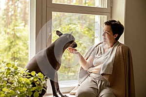 Middle age beautiful woman sitting on windowsill with her dog. 50-year-old woman spending time with her Mexican hairless