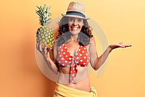 Middle age beautiful woman wearing bikini and summer hat holding pineappel celebrating achievement with happy smile and winner