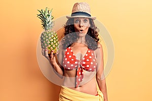 Middle age beautiful woman wearing bikini and summer hat holding pineappel scared and amazed with open mouth for surprise,
