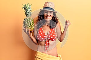 Middle age beautiful woman wearing bikini and summer hat holding pineappel screaming proud, celebrating victory and success very