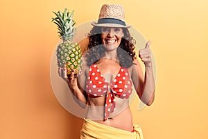 Middle age beautiful woman wearing bikini and summer hat holding pineappel smiling happy and positive, thumb up doing excellent