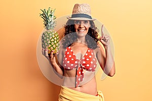 Middle age beautiful woman wearing bikini and summer hat holding pineappel smiling with an idea or question pointing finger with