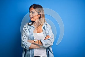 Middle age beautiful woman wearing casual shirt standing over isolated blue background looking to the side with arms crossed