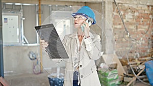 Middle age blonde woman architect talking on smartphone reading document at construction site