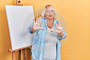 Middle age blonde woman standing by painter easel stand afraid and terrified with fear expression stop gesture with hands,