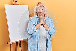 Middle age blonde woman standing by painter easel stand begging and praying with hands together with hope expression on face very