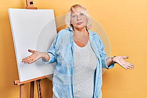 Middle age blonde woman standing by painter easel stand clueless and confused expression with arms and hands raised