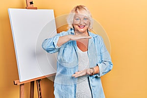 Middle age blonde woman standing by painter easel stand gesturing with hands showing big and large size sign, measure symbol