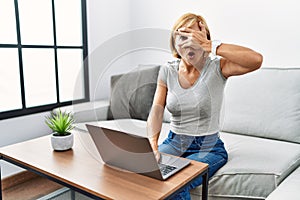 Middle age blonde woman using laptop at home peeking in shock covering face and eyes with hand, looking through fingers with