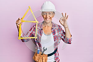 Middle age blonde woman wearing architect hardhat holding build project doing ok sign with fingers, smiling friendly gesturing