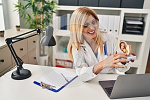 Middle age blonde woman wearing doctor uniform holding anatomical model of uterus with fetus at clinic