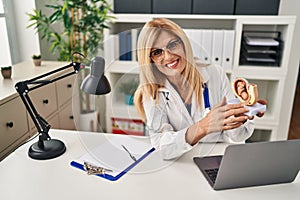 Middle age blonde woman wearing doctor uniform holding anatomical model of uterus with fetus at clinic