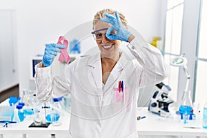 Middle age blonde woman working at laboratory looking for breast cancer cure smiling happy doing ok sign with hand on eye looking