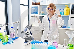 Middle age blonde woman working at scientist laboratory confused and annoyed with open palm showing copy space and pointing finger