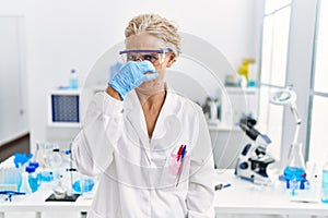 Middle age blonde woman working at scientist laboratory smelling something stinky and disgusting, intolerable smell, holding