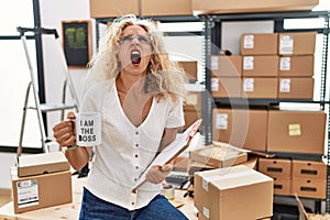 Middle age blonde woman working at small business drinking coffee cup angry and mad screaming frustrated and furious, shouting