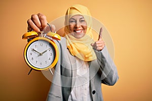 Middle age brunette business woman wearing muslim traditional hijab holding alarm clock surprised with an idea or question