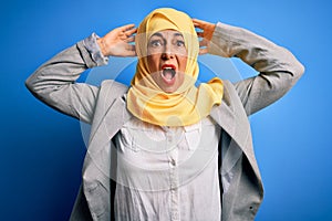 Middle age brunette business woman wearing muslim traditional hijab over blue background Crazy and scared with hands on head,