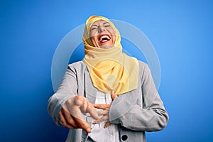 Middle age brunette business woman wearing muslim traditional hijab over blue background laughing at you, pointing finger to the